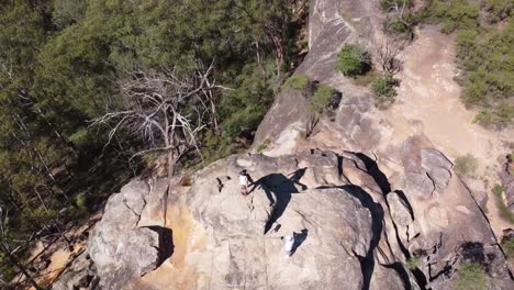 Drone-circling-a-massive-rock-in-a-forest-with-two-people-on-top-of-the-mountain