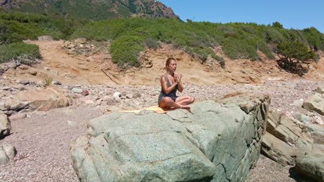Beautiful-Woman-Meditating-With-Eyes-Closed-And-Sitting-On-A-Rock-By-The-Sea-In-Sardinia,-Italy---aerial-pullback