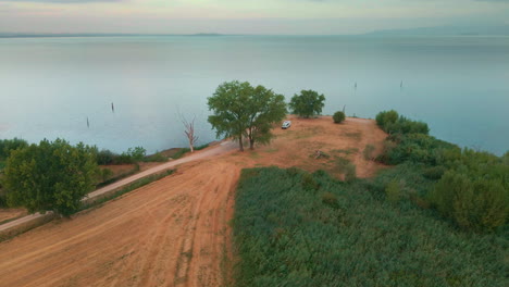 White-Camper-Van-Parked-At-The-Tip-Of-Lakeshore-During-Sunrise-With-Calm-Waters-Of-Lake-Trasimeno-In-Italy