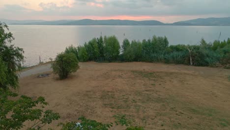 Scenic-Sunrise-At-Lake-Trasimeno-With-Calm-Waters-And-Green-Vegetation---aerial-drone-shot
