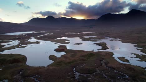 Aerial-drone-footage-reversing-above-a-peatland-landscape-of-peat-bogs-amongst-a-mosaic-of-freshwater-lochs-looking-towards-dark-mountains-on-the-horizon-at-sunset
