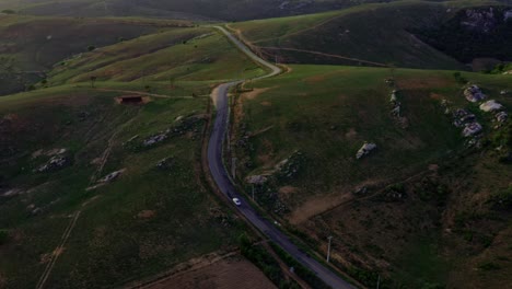 Drone-shot-of-a-beautiful-windy-road-surrounded-by-hills,-rocks,-and-farmland-with-a-small-car-drying-down-it-in-the-countryside-of-Rio-Grande-do-Norte-in-the-Northeast-of-Brazil-during-golden-hour
