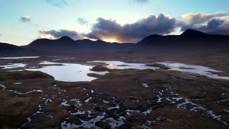 Drone-footage-flying-above-of-a-patchwork-wetland-landscape-of-islands-and-peat-bogs-surrounded-by-fresh-water-looking-towards-dark-mountains-on-the-horizon-at-sunset