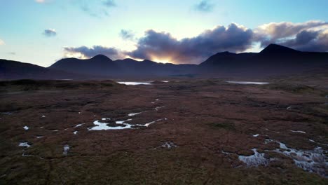 Aerial-drone-footage-flying-low-and-close-to-the-surface-of-a-peat-bogs-before-rising-to-reveal-freshwater-lochs-and-dark-mountains-on-the-horizon-at-sunset