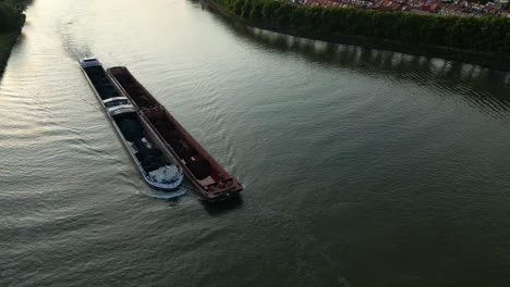 AERIAL---Two-cargo-barges-in-a-river-in-The-Netherlands,-wide-reverse-shot