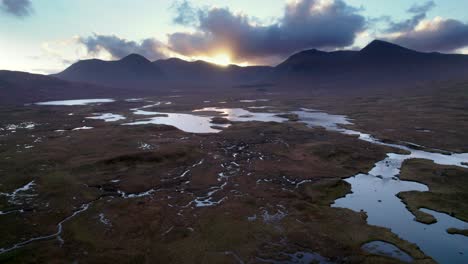 A-drone-slowly-descends-towards-a-wetland-landscape-of-islands-and-peat-bogs-surrounded-by-fresh-water-while-looking-towards-dark-mountains-at-sunset