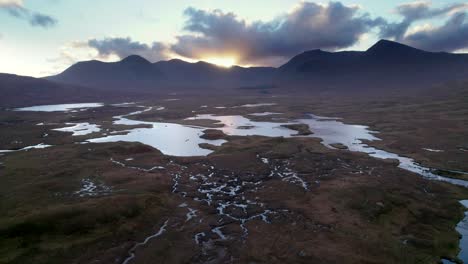 A-drone-slowly-rolling-above-a-wetland-landscape-of-islands-and-peat-bogs-surrounded-by-fresh-water-while-looking-towards-dark-mountains-at-sunset