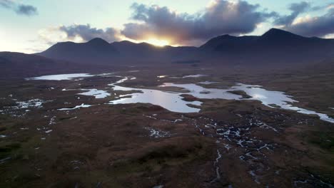 A-drone-reverses-high-above-a-peatland-landscape-of-peat-bogs-amongst-a-mosaic-of-fresh-water-lochs-looking-towards-dark-mountains-on-the-horizon-at-sunset
