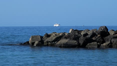 Stone-pier-at-the-beach-of-Playa-De-Las-Americas-,-sunny-summer-day,-calm-Atlantic-ocean-in-the-background,-distant-tourist-ferry,-medium-handheld-shot