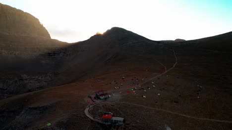wide-aerial-shot-of-a-valley-under-the-peak-of-mountain-Olympus-while-the-sun-sets-and-creates-lens-flares,-the-shot-continues-and-lowers-altitude-revealing-a-camper's-hut-and-lots-of-colourful-tens