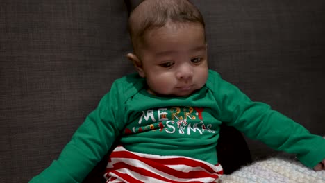 Adorable-Bangladeshi-Baby-Wearing-Festive-Green-Merry-Christmas-Outfit-Sat-On-Sofa-Looking-Side-To-Side