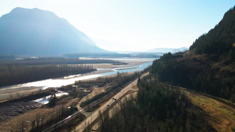 Aerial-Shot-of-the-Fraser-River-next-to-the-Lougheed-Highway-7-in-the-Lower-Mainland-in-British-Columbia-in-Canada,-bright-scene-with-foggy-mountains-in-the-autumn-with-cars-driving-on-the-road