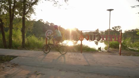 A-stationary-panning-footage-of-a-commuting-woman-riding-her-bike-with-panniers-passing-by-a-public-park-at-the-riverside-in-Isan,-Thailand