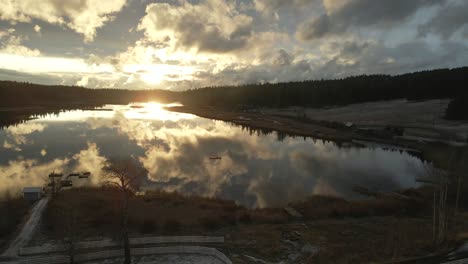 Magnificent-Aerial-Scene-of-Glimpse-Lake-during-sunset-in-golden-hour-light,-on-a-cloudy-day,-beautiful-reflections-of-the-sky-in-the-water,-reverse-flight-over-ranch-land-with-majestic-spruce-trees