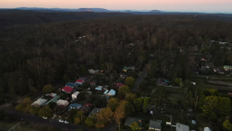 High-lowering-Drone-shot-over-town-with-road-and-traffic-surrounded-by-dense-forest-bushland