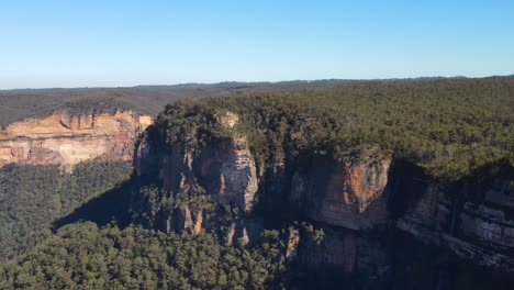 Dramatic-aerial-drone-flyover-of-rock-outcrop-covered-in-trees-and-bushes-in-the-Blue-Mountains-clear-blue-sky-in-New-South-Wales
