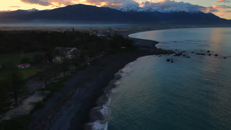 Aerial-drone-shot-of-sea-with-lapping-waves-and-snow-capped-mountain-background-houses-orange-dusk-sky-new-Zealand