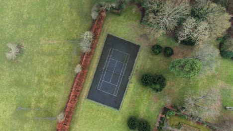 Top-down-drone-view-of-private-asphalt-tennis-court-in-garden-surrounded-by-trees,-grass-and-fields
