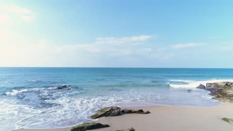 Flying-forward-over-a-rocky-sand-beach-towards-the-blue-azure-sea-water-in-Cadiz,-Spain