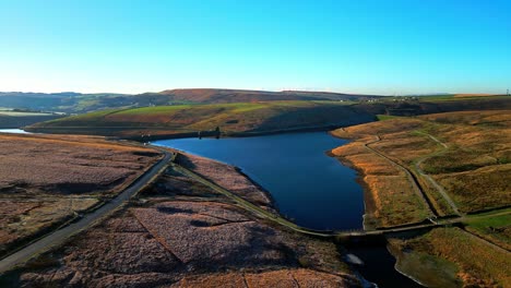 Sweeping-aerial-footage-flying-over-golden-moorlands-scattered-with-frost-directley-towards-a-lake,-reservoir-in-the-distance