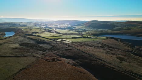 Low-sweeping-aerial-footage-flying-into-the-sun-and-over-golden-moorlands-scattered-with-frost-directley-towards-a-lake,-reservoir-in-the-distance