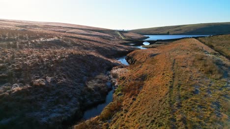 Aerial-footage-flying-over-golden-moorlands-and-over-a-small-river,-stream-directley-towards-a-stone-built-bridge-and-lake,-reservoir-Hills,-valley,-and-blue-sky