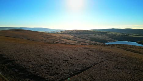 Winter-aerial-footage-flying-over-golden-moorlands-directley-into-the-sun-with-lake,-reservoir-in-the-distance