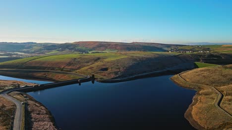 Low-sweeping-aerial-footage-flying-over-golden-moorlands-scattered-with-frost-directley-towards-a-lake,-reservoir-in-the-distance