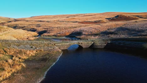 Winter-aerial-footage-of-a-small-stone-built-bridge-crossing-a-reservoir-and-river-high-on-the-Pennine-hills,-West-Yorkshire,-England