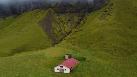 Single-lonely-house-at-base-of-the-mountain-in-Icelands-countryside-with-low-clouds-and-rolling-meadows-around,-remote-summer-cottage