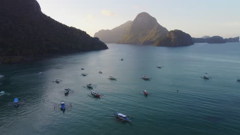 Flying-Over-Anchored-Boats-In-Front-Of-High-Distant-Mountains,-El-Nido,-Philippines