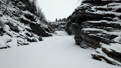 Drone-flying-over-frozen-river-between-rocky-riverbanks-in-northern-Norway,-Scandinavia