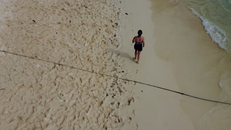 Drone-shot-of-a-girl-walking-along-shoreline-of-a-white-sand-beach