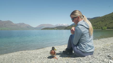 Attractive-blond-woman-feeding-bread-to-duck-while-sitting-on-pebble-beach