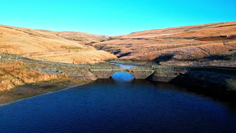 Aerial-video-footage-of-a-small-stone-built-bridge-crossing-a-reservoir-and-river-high-on-the-Pennine-hills,-West-Yorkshire,-England