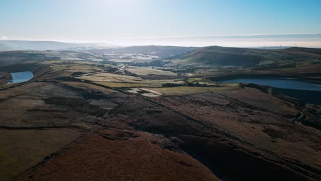 Low-sweeping-aerial-footage-flying-into-the-midday-sun-and-over-golden-moorlands-scattered-with-frost-directley-towards-a-lake,-reservoir-in-the-distance