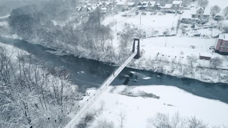 Aerial-view-of-old-hanging-bridge-over-frozen-river-in-frosty-snowy-morning