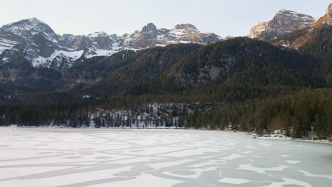 Icy-Lake-With-Dense-Forest-Trees-And-Snowcapped-Mountain-Range-Of-Dolomites-In-Trentino,-Italy