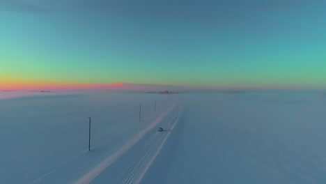 Car-driving-down-a-snow-covered-countryside-road-at-twilight---aerial-follow