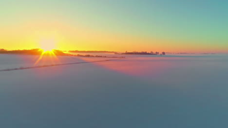 Glowing-golden-sunset-on-the-horizon-of-an-empty-snowy-landscape-of-rural-farmland-fields---aerial-view