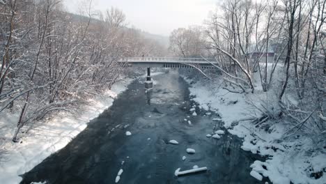 Bird-eye-view-of-old-bridge,-frozen-river-and-fallen-trees-covered-by-snow