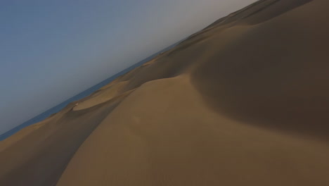 Aerial-fast-low-flight-over-sand-dunes-towards-sea-at-sunset,-Maspalomas