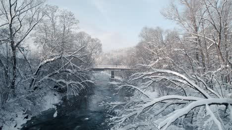 Drone-view-of-frozen-river,-snowy-broken-trees-and-bridge-surrounded-by-forest