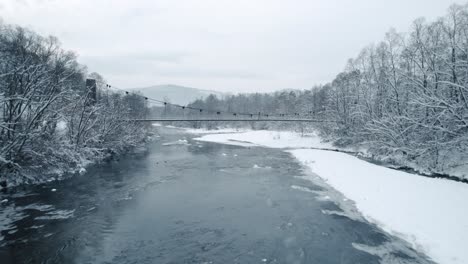 Luftaufnahme-Des-Zugefrorenen-Flusses-Mit-Schwimmenden-Enten,-Hängebrücke-Und-Wandermann-In-Der-Wintersaison
