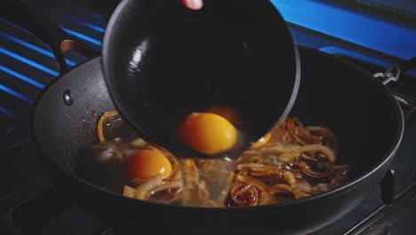 Adding-eggs-to-a-frying-pan-with-fried-onion-on-it,-close-up-view