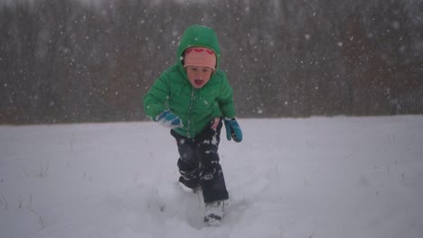 Kleiner-Junge-Rennt-Und-Taucht-In-Den-Schnee,-Lächelt-Dann-In-Die-Kamera,-Fallender-Schnee-In-Zeitlupe