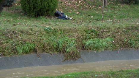 Obdachloser-Bettler-Schläft-Am-Ufer-Des-Schmutzigen-Wasserkanals-Der-Stadt,-Der-Nach-Heftigen-Regen-Fließt