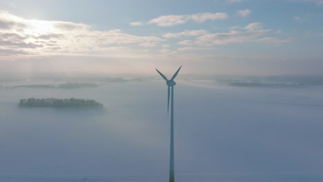 Establishing-aerial-view-of-wind-turbine-generating-renewable-energy-in-the-wind-farm,-snow-filled-countryside-landscape-with-fog,-sunny-winter-day,-wide-drone-shot-moving-forward