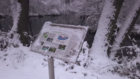 Snow-covered-information-board-overlooking-Niebieskie-Zrodla-nature-reserve-woodland-river,-pull-back-shot