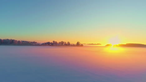 Aerial-drone-shot-over-frozen-lake-during-cold-winter-foggy-sunrise-with-silhouette-of-trees-visible-in-the-background